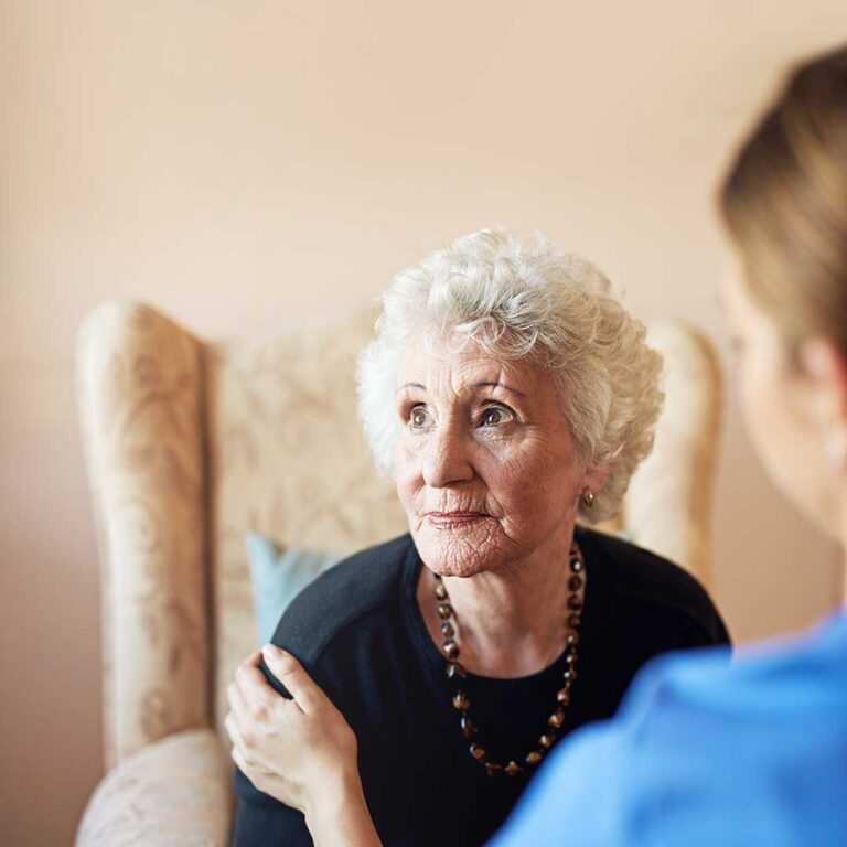 Older resident uses a tablet at a table while a caregiver reviews a safety alert device.